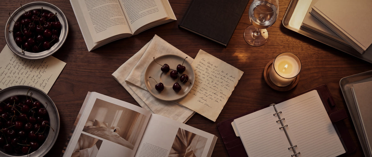 A walnut table at night with cherries, handwritten notes, an open book and a lit candle