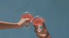 Two people raise glasses of Glow State by UMII, a soft rose-pink daily wellness drink, in a toast against a teal wall.