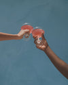 Two people raise glasses of Glow State by UMII, a soft rose-pink daily wellness drink, in a toast against a teal wall.